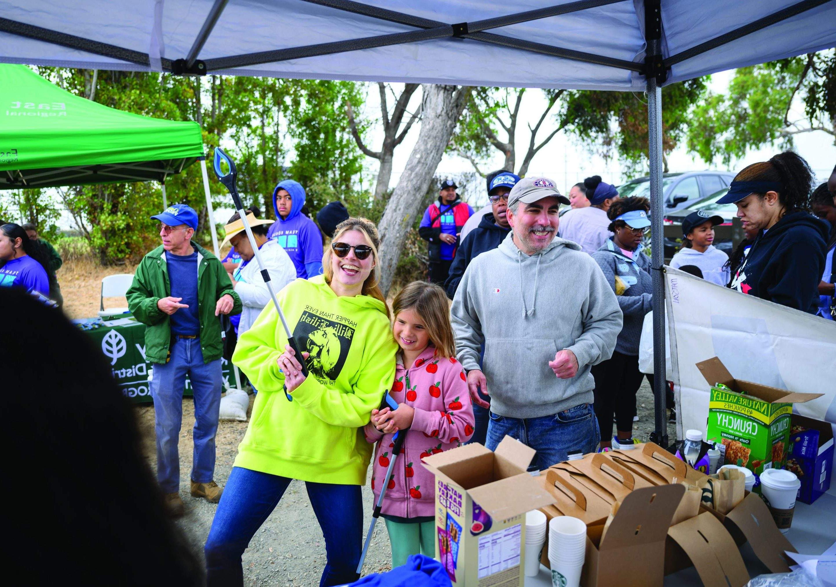 Family stands near table of snacks with other volunteers and pop-up tents in the background.