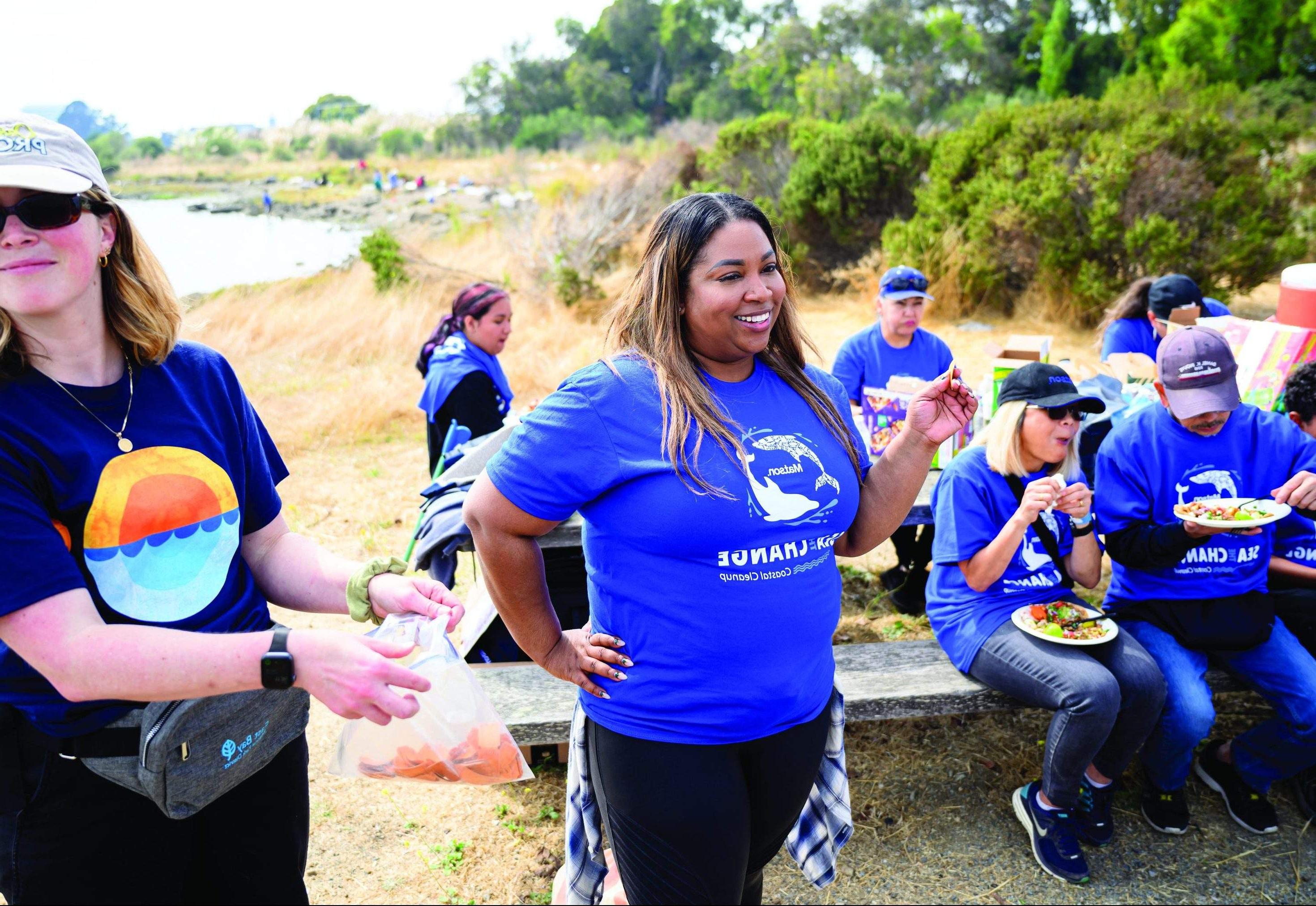 Denise stands with a raffle ticket in hands as volunteers wearing blue 澳博体育app下载 "Sea The Change" t-shirts sit on a picnic table behind her eating.